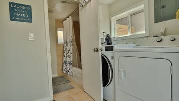 A laundry room with a sign reading "Laundry today or naked tomorrow," a washing machine, a dryer, a window, and a shower area with a curtain.