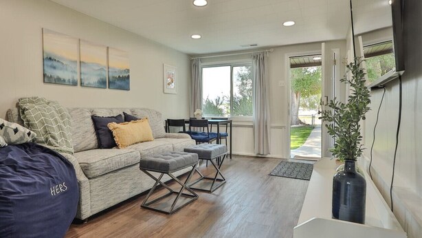 A modern living room with a sofa, ottomans, a dining table, and chairs. There is a large window with curtains, a plant on a side table, and artwork on the wall. - Courtyard Hot Tub in the Heart of Downtown (Panguitch)