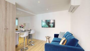 Un salon moderne avec un canapé bleu, des chaises blanches et un coin cuisine à l’arrière-plan.