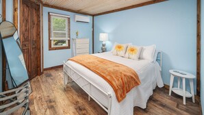 Een slaapkamer met een houten plafond, een bed met witte beddengoed, een houten dressoir, een spiegel en een klein tafeltje met een lamp.