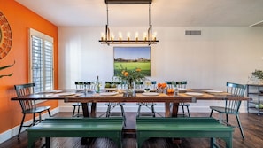 A dining area with a long table, chairs, a hanging light fixture, and a large window with blinds.