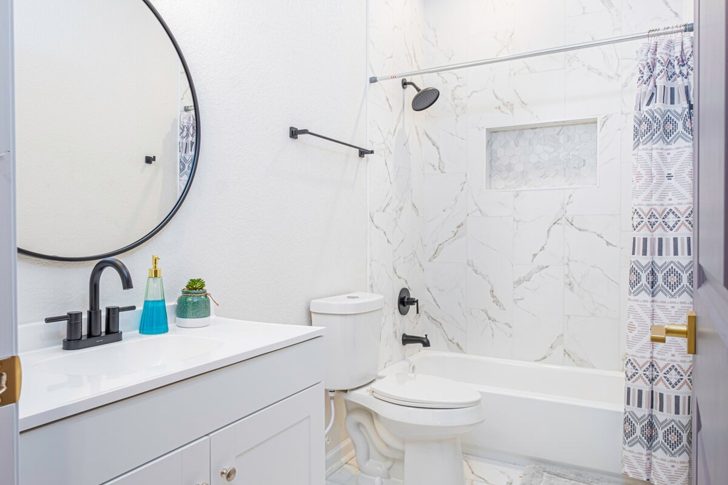 Ein modernes Badezimmer mit einer weißen Marmorwand, einem runden Spiegel, einer schwarzen Armatur, einer weißen Toilette, einem weißen Waschtisch und einer Dusche mit Handbrause sowie einem waschbunt gemusterten Duschvorhang.