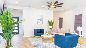 Una sala de estar moderna con ventilador de techo, una planta grande en maceta, un sofá color beige, sillones azules y una mesa de centro.