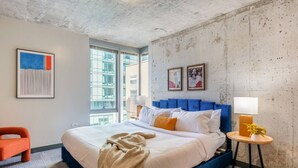 Habitació d'hotel moderna amb un llit gran, un panell blau al capçal, una petita taula auxiliar amb una làmpada i vista a la ciutat a través de grans finestres.
