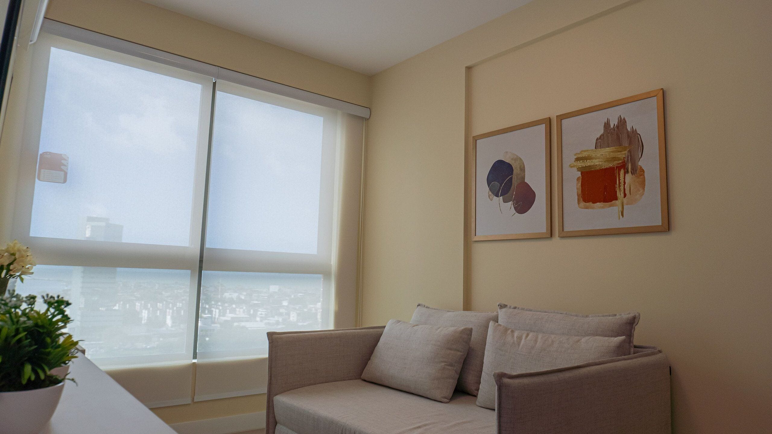 A living room with a sofa, a window with blinds, and two framed artworks on the wall.