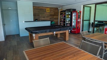 Una mesa de billar con tela negra, rodeada de sillas de madera y una mesa. Hay dos máquinas expendedoras, una de Coca-Cola y otra con un logotipo amarillo. En el fondo se ve un mostrador de madera con el logotipo de una cesta.