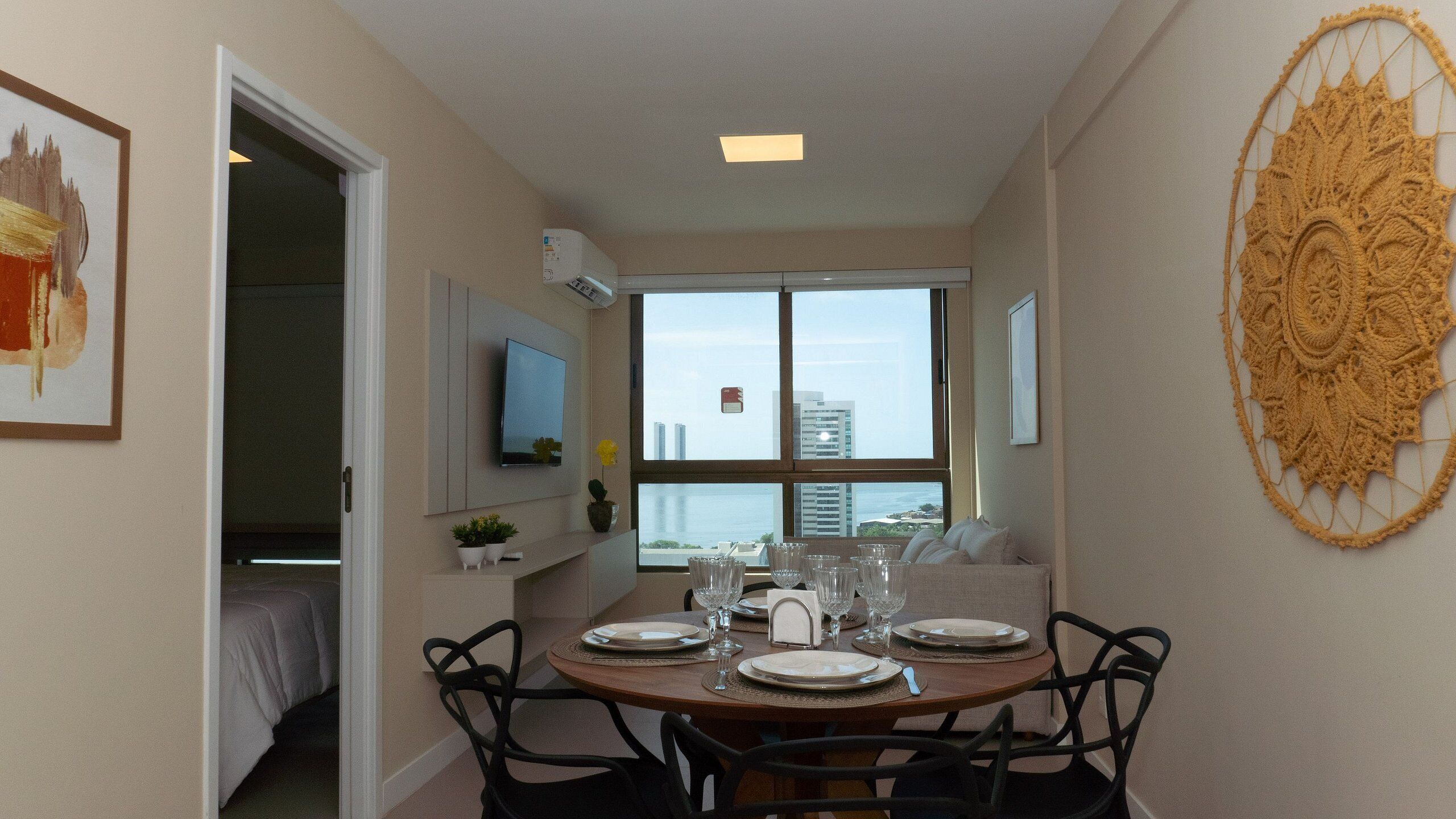 A dining area with a round table set for four, a television mounted on the wall, and a large window offering a view of buildings and the sky.