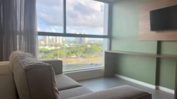 A modern living room with a sofa facing a large window, a flat-screen TV mounted on the wall, and a view of a cityscape.