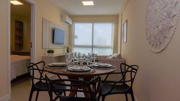 A dining area with a round table set for four, surrounded by black chairs. There is a television mounted on the wall, a window with a view of the city, and a decorative lace piece on the wall.