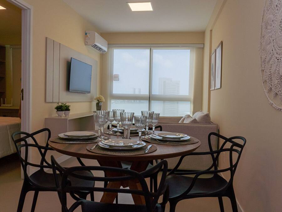 A dining area with a round table set for four, surrounded by black chairs. There is a television mounted on the wall, a window with a view of the city, and a decorative lace piece on the wall.