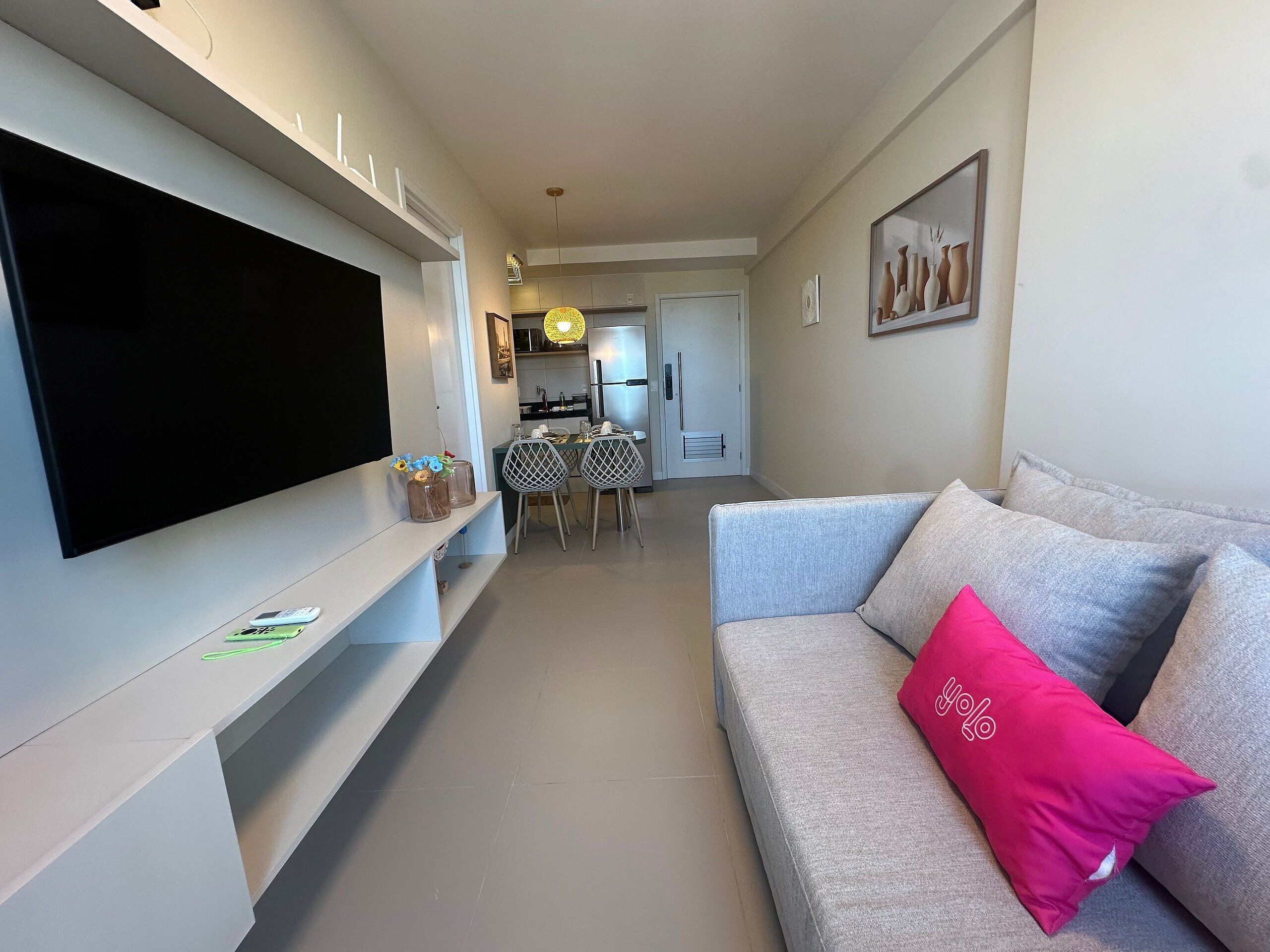 Una sala de estar con un televisor de pantalla plana, un sofá gris con un cojín rosa, una zona de comedor con mesa y sillas, y una cocina al fondo.