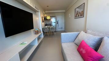 Una sala de estar con un televisor de pantalla plana, un sofá gris con un cojín rosa, una zona de comedor con mesa y sillas, y una cocina al fondo.