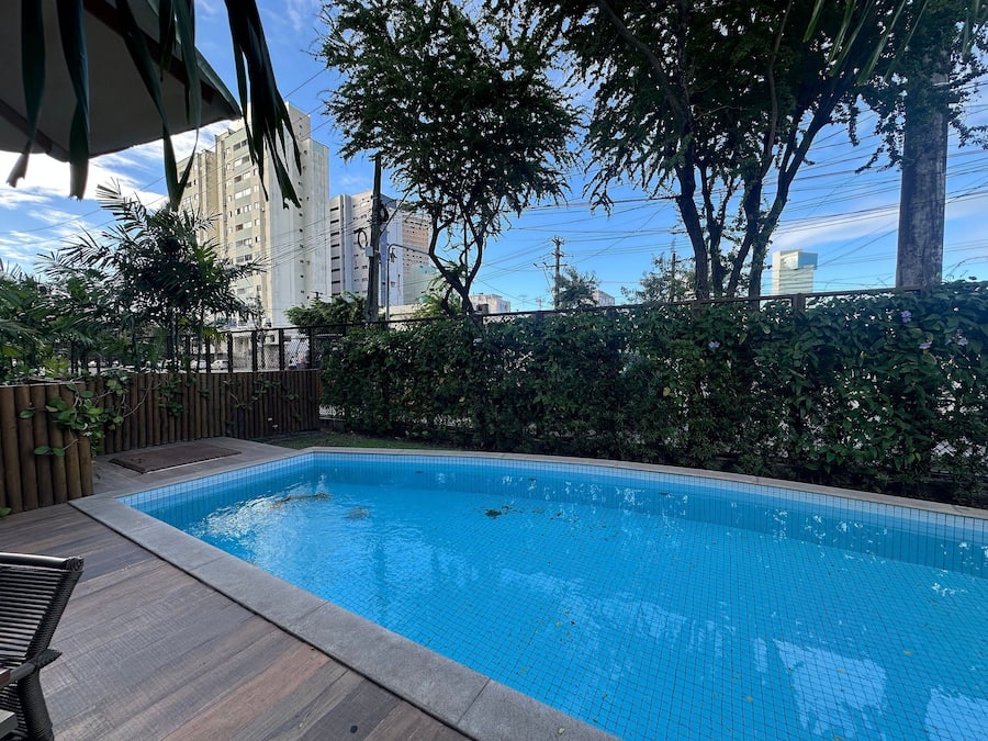 Una piscina con suelo de baldosas, rodeada de vegetación y una valla.