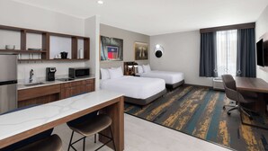 A hotel room with a kitchenette, two beds, a desk, and a TV.