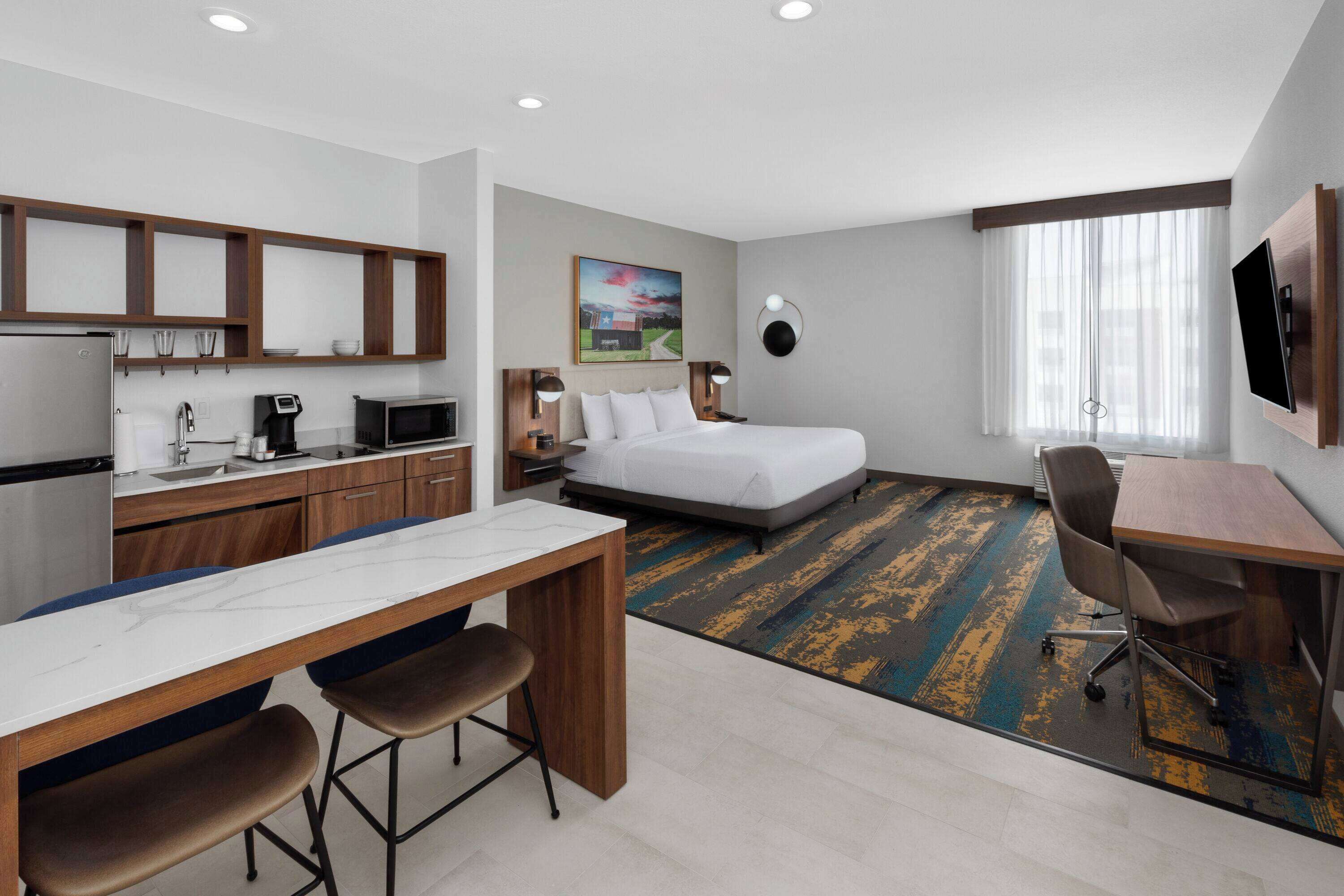 A hotel room with a bed, a desk with two chairs, a kitchenette, a TV, and a painting on the wall.