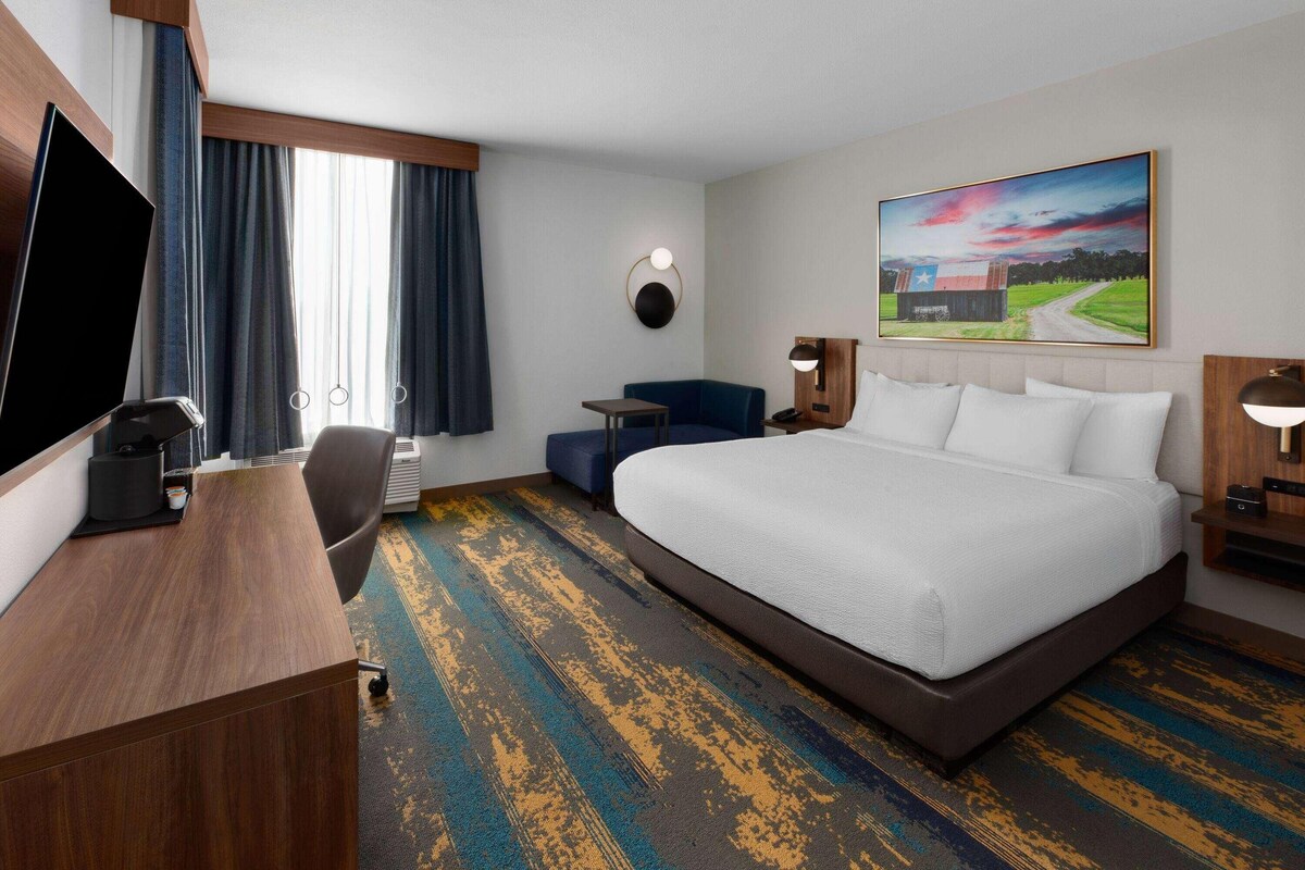 A hotel room with a bed, a desk, a chair, a TV, and a painting on the wall.