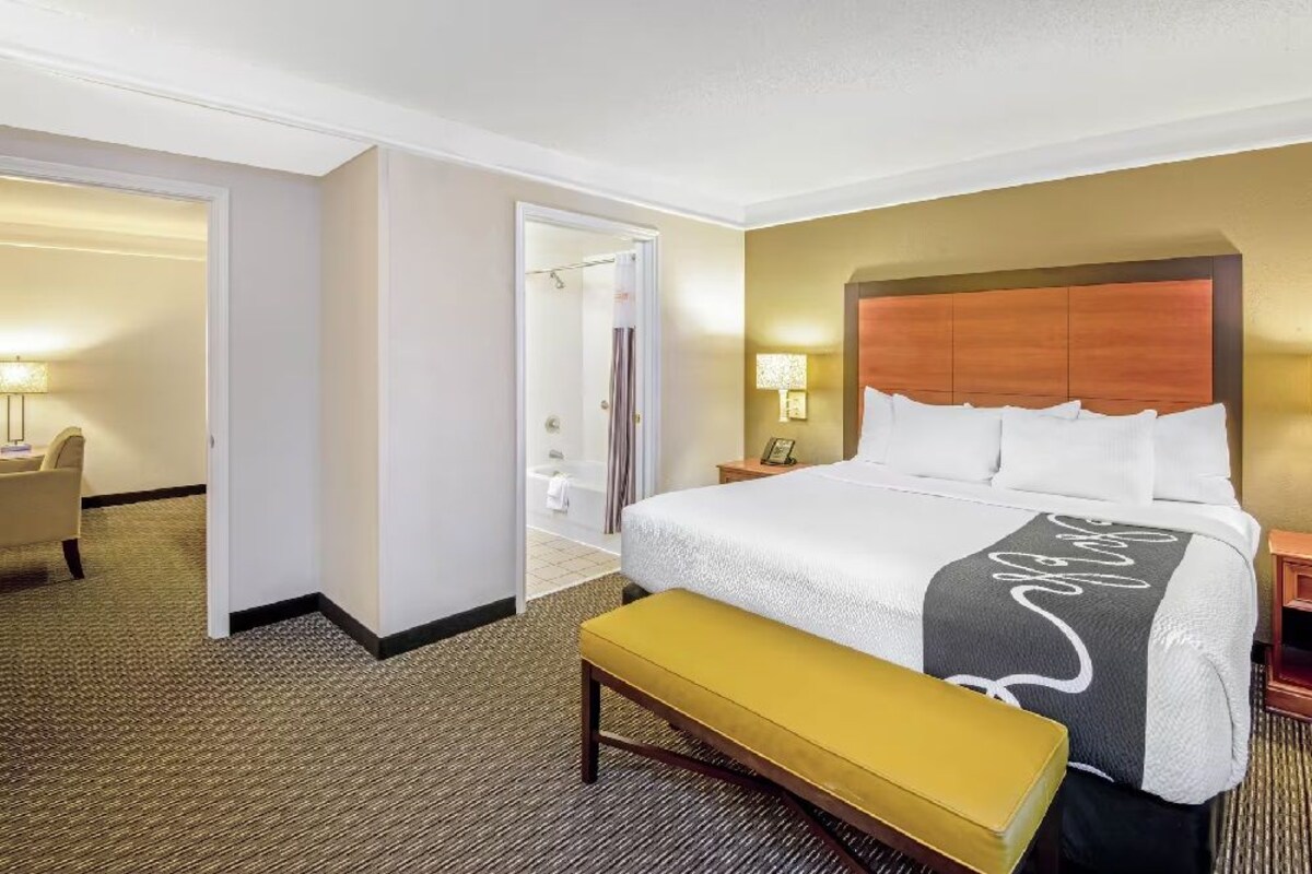 A hotel room with a large bed, a bench, a nightstand, a lamp, and a bathroom visible through an open door.