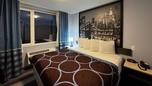 Habitació d'hotel amb un llit, una finestra amb cortines, una tauleta de nit amb un telèfon i una obra d'art mural amb una vista urbana.