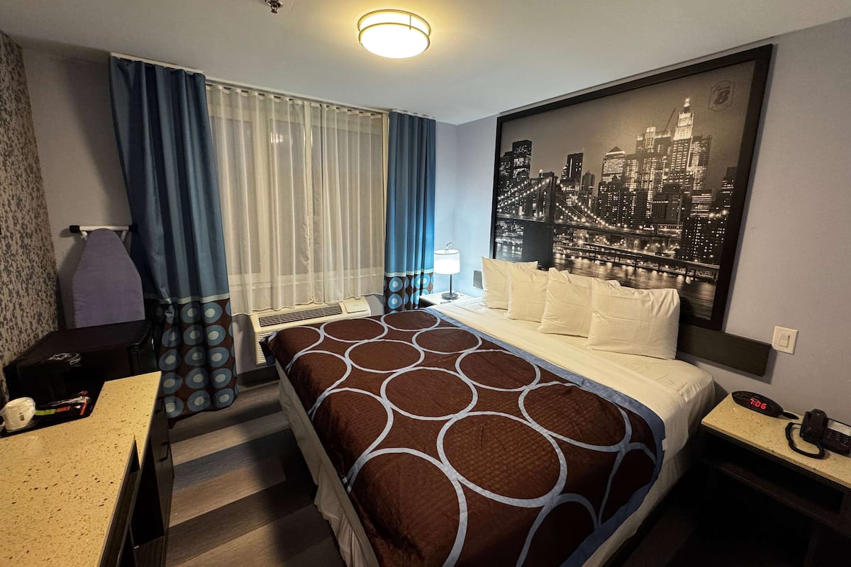 Habitación de hotel con cama, escritorio, silla, mesita de noche, reloj y ventana con cortinas.