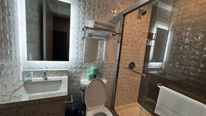 Bany amb un pica de marbre, un vàter, una cabina de dutxa de vidre i un suport per a tovalloles.