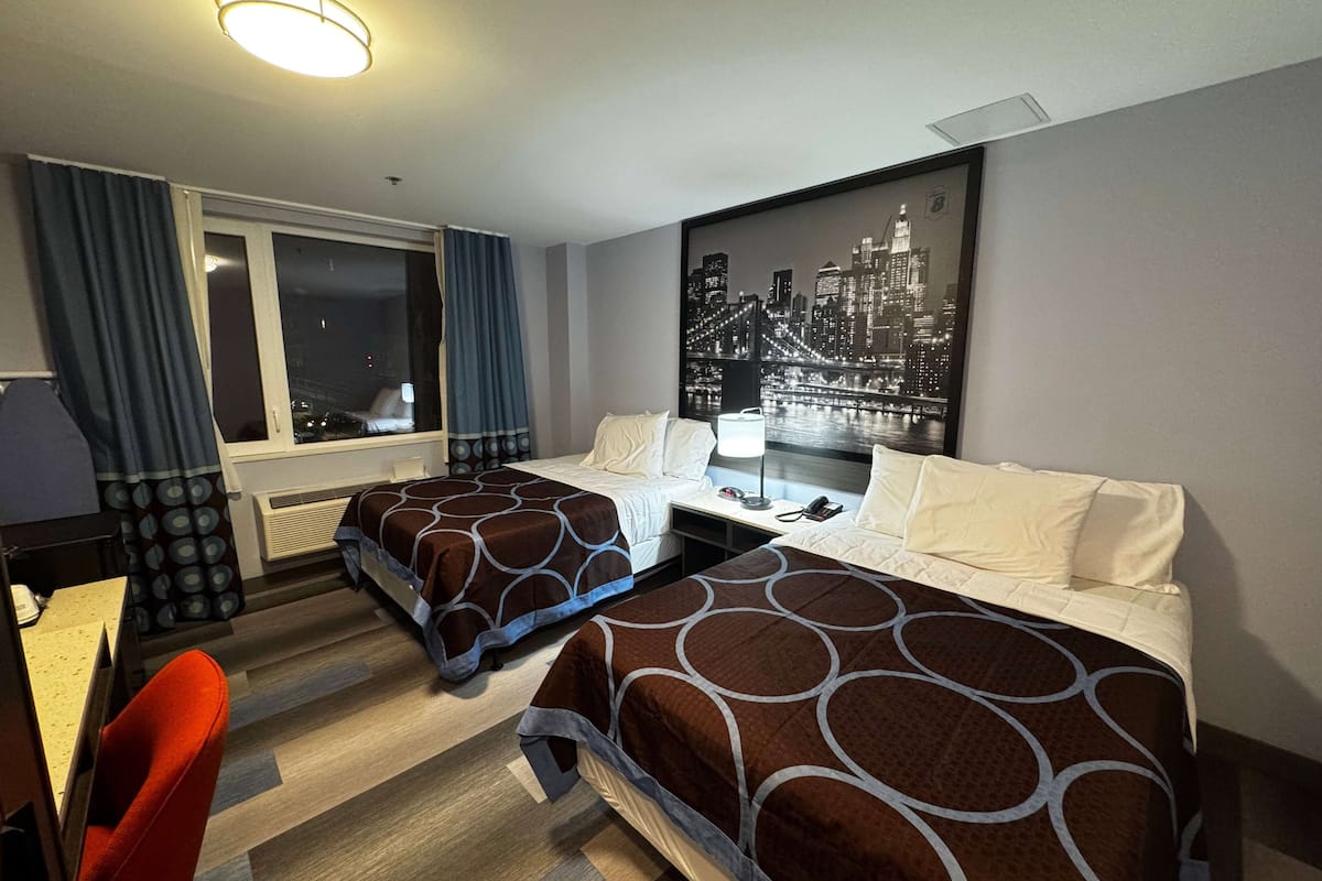 Habitación de hotel con dos camas, un escritorio, una silla, un ventanal con cortinas y una obra de arte con un paisaje urbano en la pared.