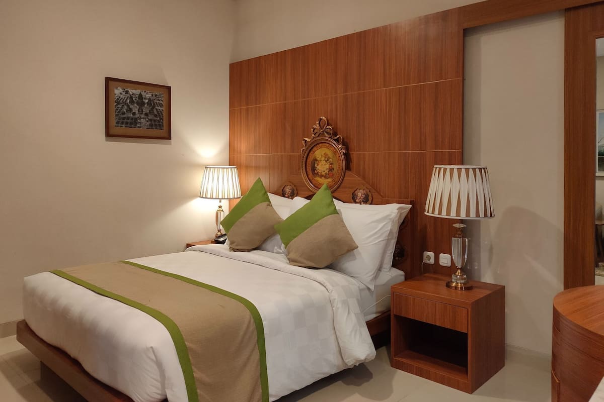 Una cama bien tendida con colcha verde y beige, dos lámparas de noche y un cabecero de madera con un espejo decorativo.