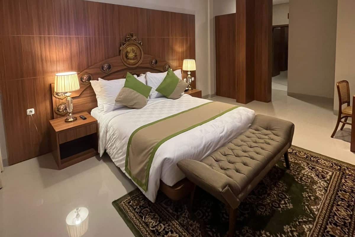 Un dormitorio moderno con una cama grande, cabecera de madera, dos mesitas de noche con lámparas, un banco y una alfombra estampada.