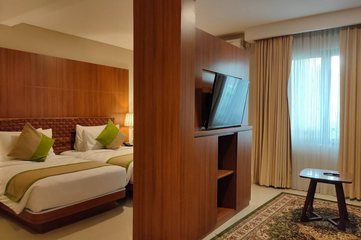 Habitación de hotel con cabecera de madera, televisor de pantalla plana, cama con ropa blanca y almohadas verdes, una mesita pequeña y un ventanal grande con cortinas.