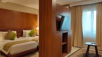 Habitación de hotel con cabecera de madera, televisor de pantalla plana, cama con ropa blanca y almohadas verdes, una mesita pequeña y un ventanal grande con cortinas.