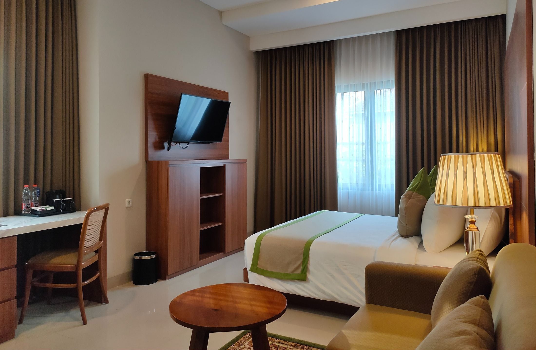 Habitación de hotel con cama, escritorio con silla, televisor en la pared, lámpara y ventana con cortinas.