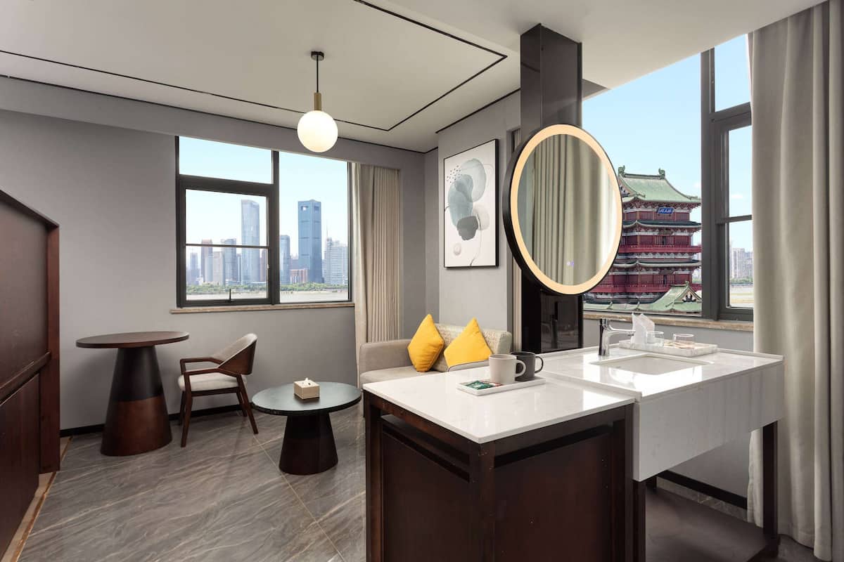 A modern hotel room with a large window offering a city view, a round mirror, a white sink, and a small round table with a chair.