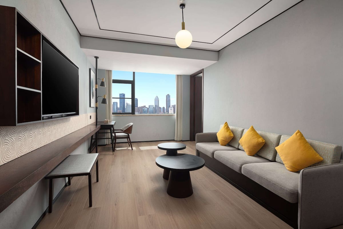 A modern living room with a flat-screen TV, a sofa with yellow cushions, a round coffee table, and a city view through large windows.