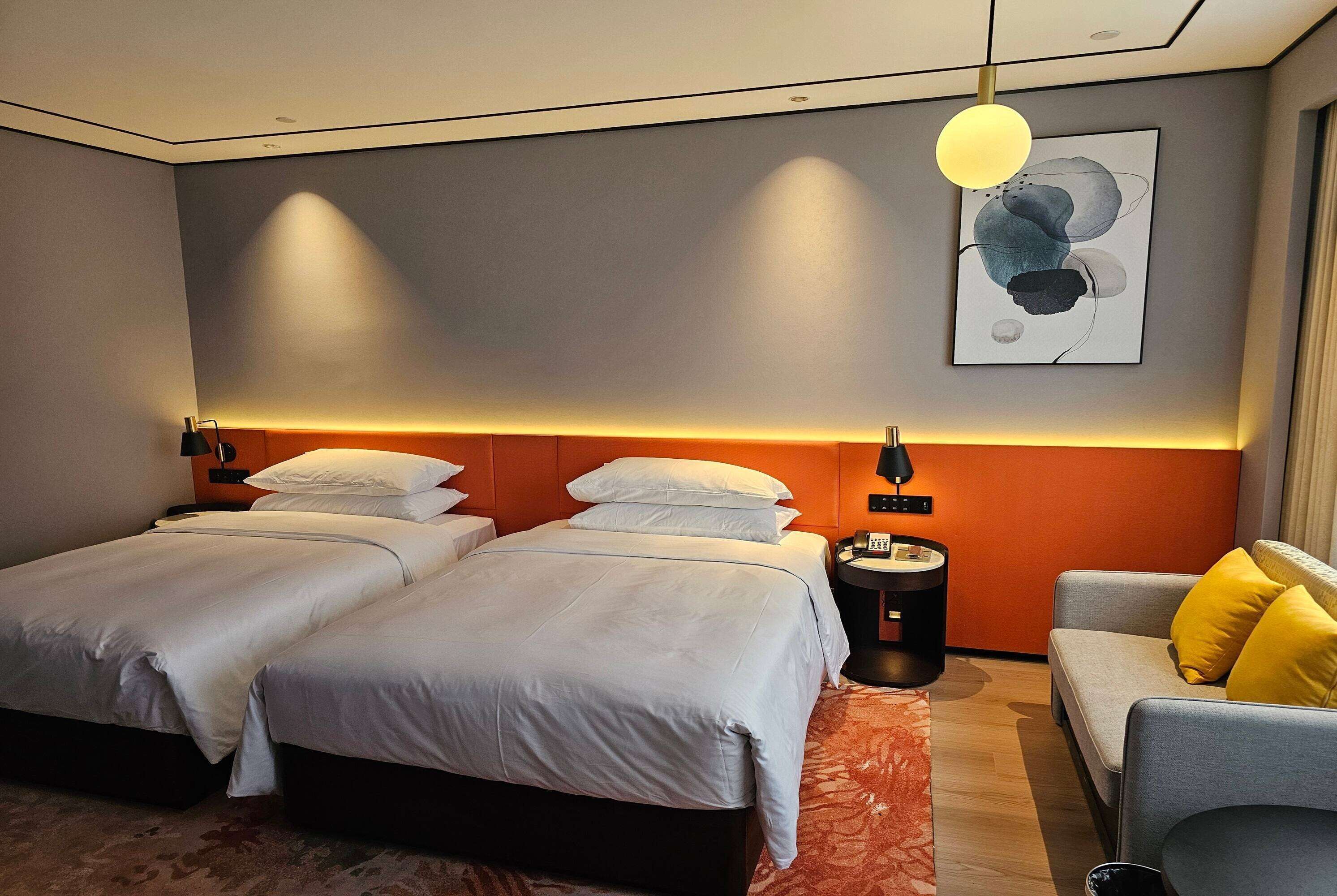 A hotel room with two beds, a sofa, a bedside table, and a wall-mounted artwork.