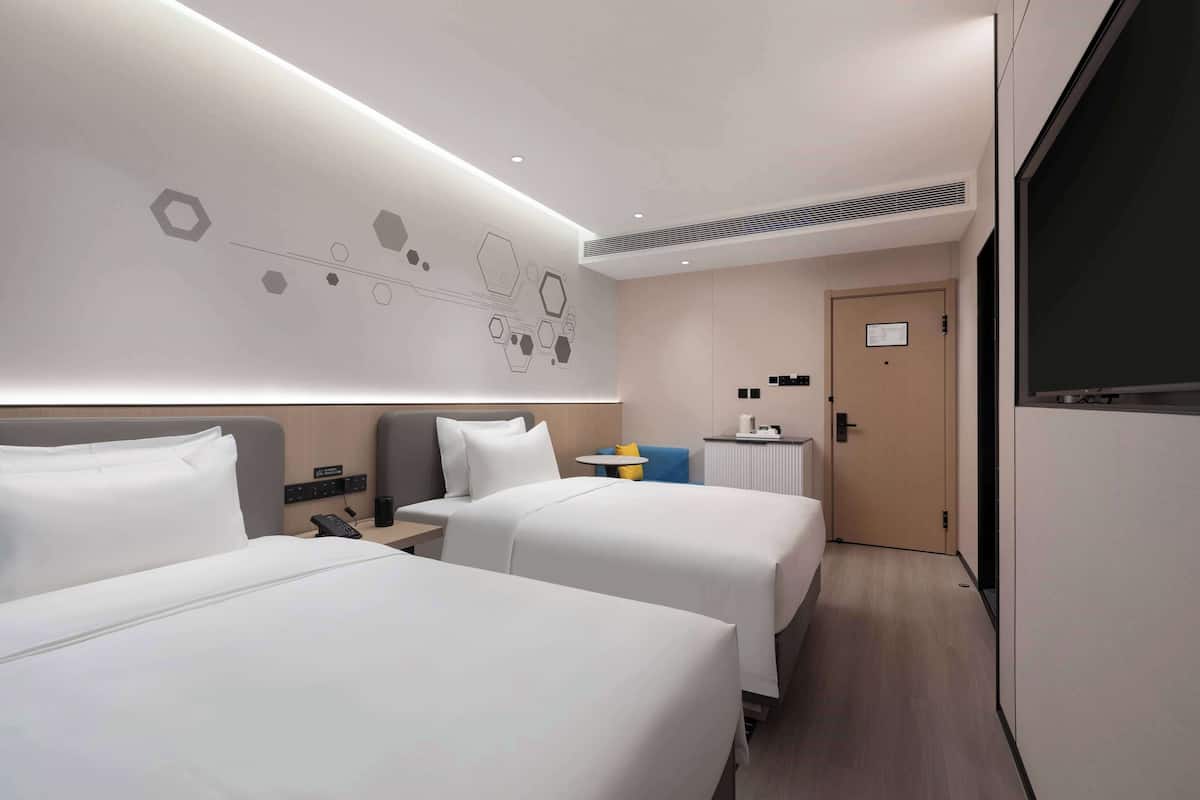 A hotel room with two beds, a bedside table with a phone, a small table with a lamp, and a door leading to another room.