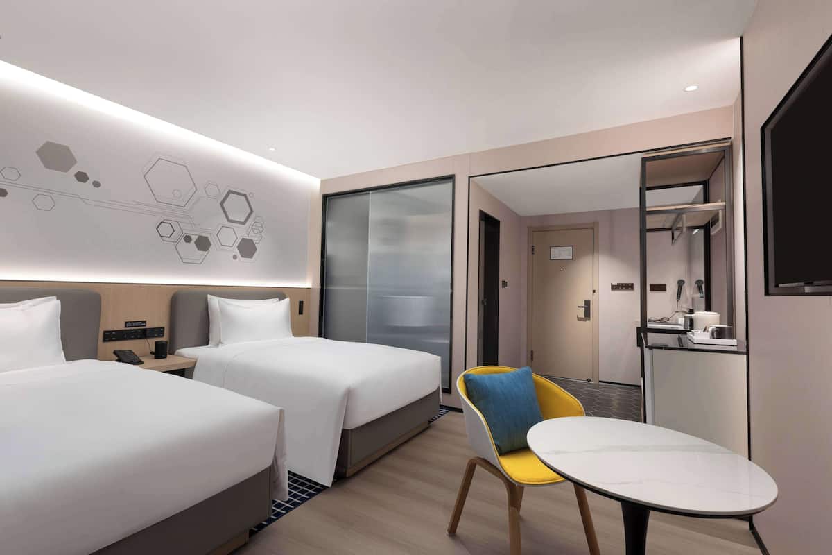A modern hotel room with two beds, a small table, a chair, and a bathroom visible through a glass door.