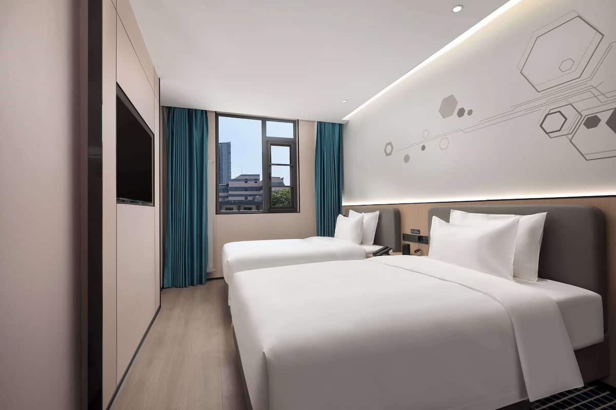 A hotel room with two beds, a flat-screen TV, a nightstand, and a view of the city through large windows.