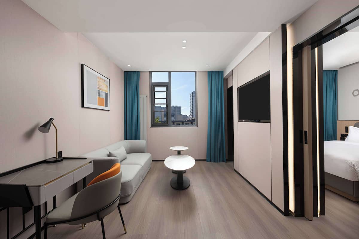A modern hotel room with a desk, sofa, two small tables, a bed, and a large window with curtains.