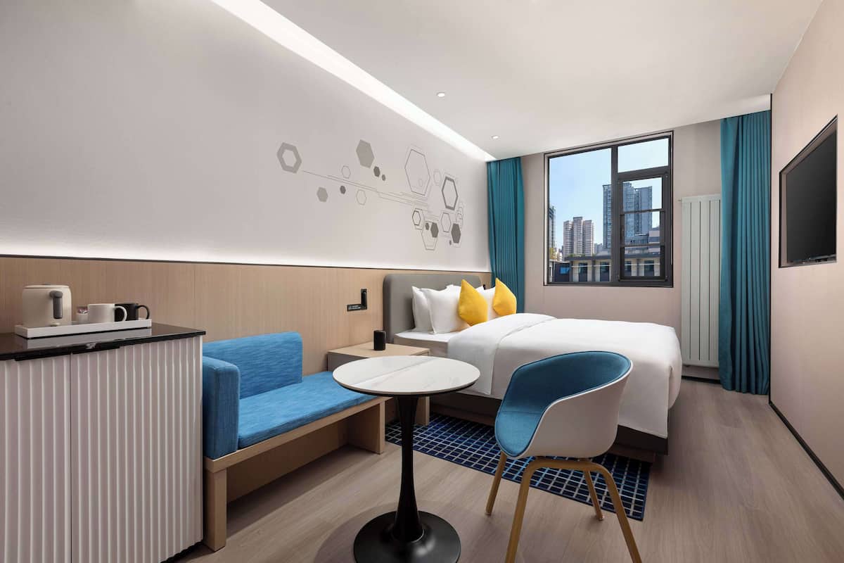 A modern hotel room with a bed, a desk with a chair, a small table, and a view of the city through large windows.