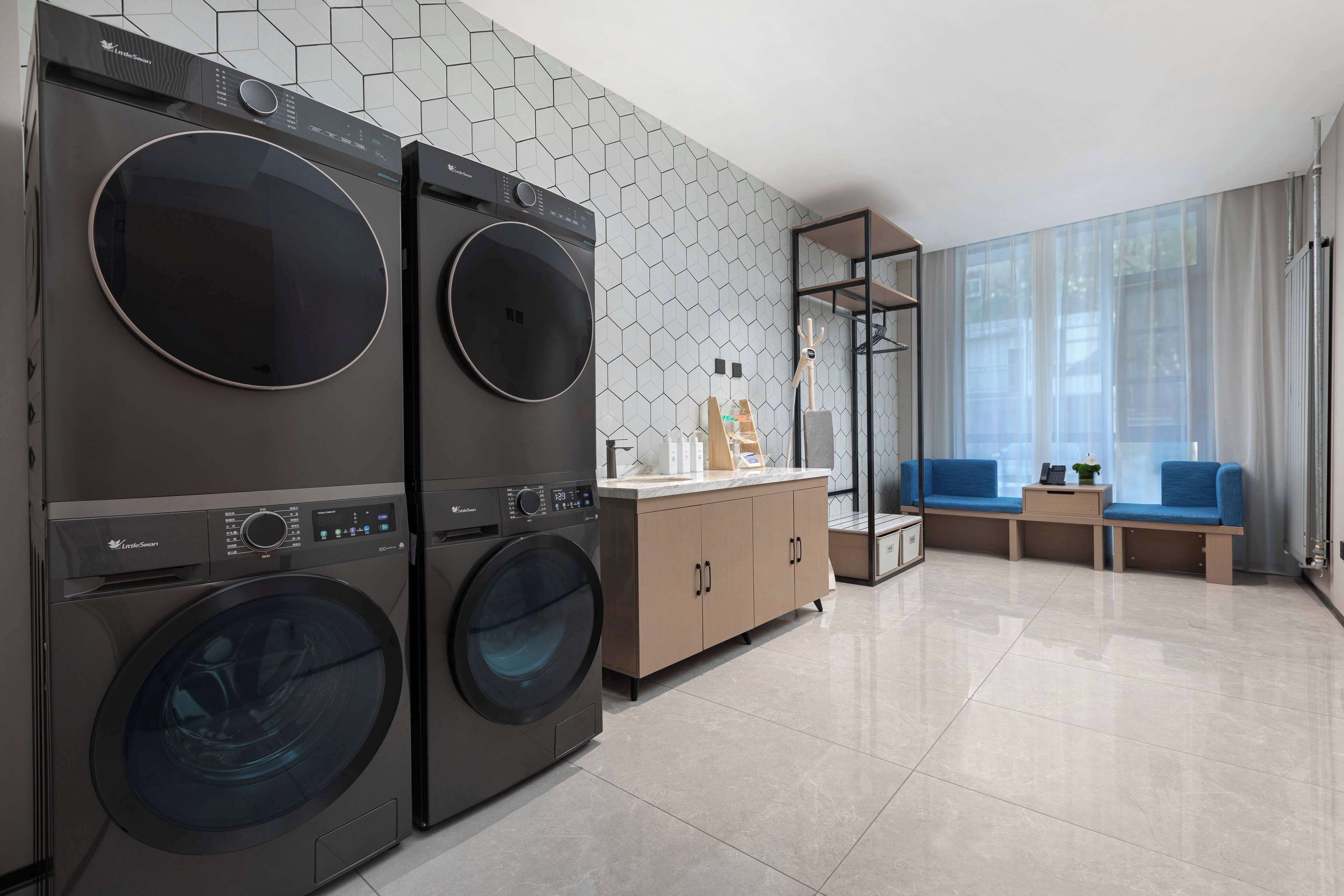 Een moderne wasruimte met twee voorladers, een houten kast, en een zithoek met een bank en een bijzettafeltje.