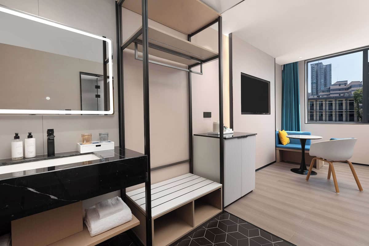 A modern hotel bathroom with a large mirror, black marble sink, and a small seating area with a table and chair.