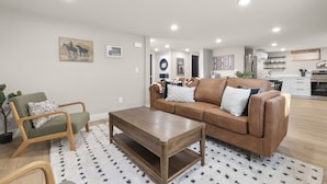 A modern living room with a brown leather sofa, a wooden coffee table, and a green armchair. There is a kitchen in the background with white cabinets and a refrigerator. - Steps to Hospital and MSU-B| Renovated Hospital Retreat (Billings)