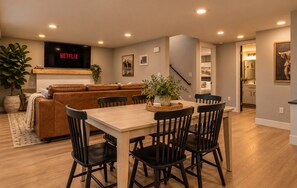 A modern living room with a wooden dining table, black chairs, a brown leather sofa, and a flat-screen TV displaying 'NETFLIX'.