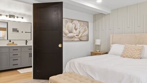 A bedroom with a large bed, a dark door, a wall-mounted mirror, and a large floral painting.