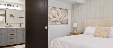 Un dormitorio con una cama grande, una puerta oscura, un espejo montado en la pared y un cuadro floral de gran tamaño.