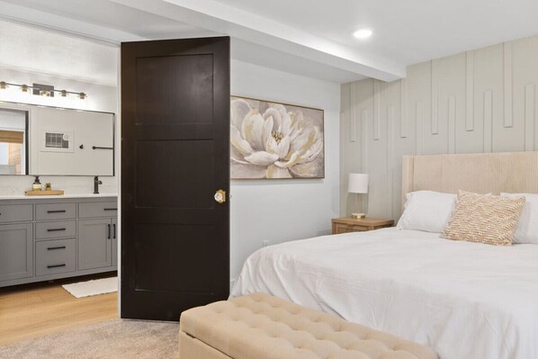 Un dormitorio con una cama grande, una puerta oscura, un espejo montado en la pared y un cuadro floral de gran tamaño.