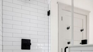 Un baño con paredes de azulejos tipo metro blancos, una puerta blanca con manija negra y un estante blanco con dos botellas.