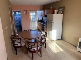 A dining area with a round wooden table and chairs, a kitchen with wooden cabinets and appliances, and a tiled floor.