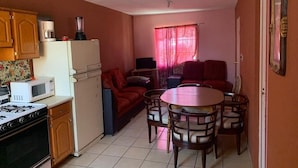 Cocina compacta con estufa, microondas y refrigerador. Hay un comedor con mesa y sillas, y un sofá al fondo.