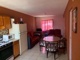 A compact kitchen with a stove, microwave, and refrigerator. There is a dining area with a table and chairs, and a sofa in the background.