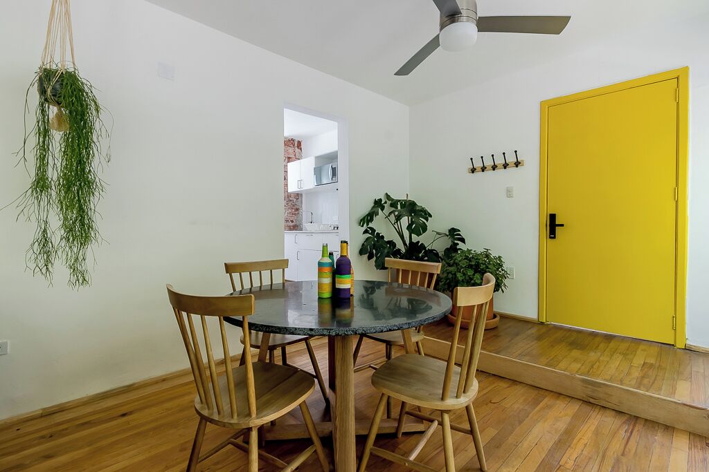 Un coin repas avec une table ronde en verre, des chaises en bois, une plante suspendue, un ventilateur de plafond et une porte jaune.